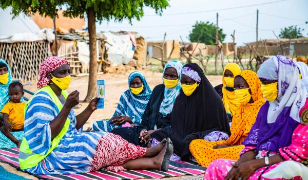 Women in colourful garments wearing yellow masks are sitting on mats outdoors, listening to a woman holding up a leaflet.