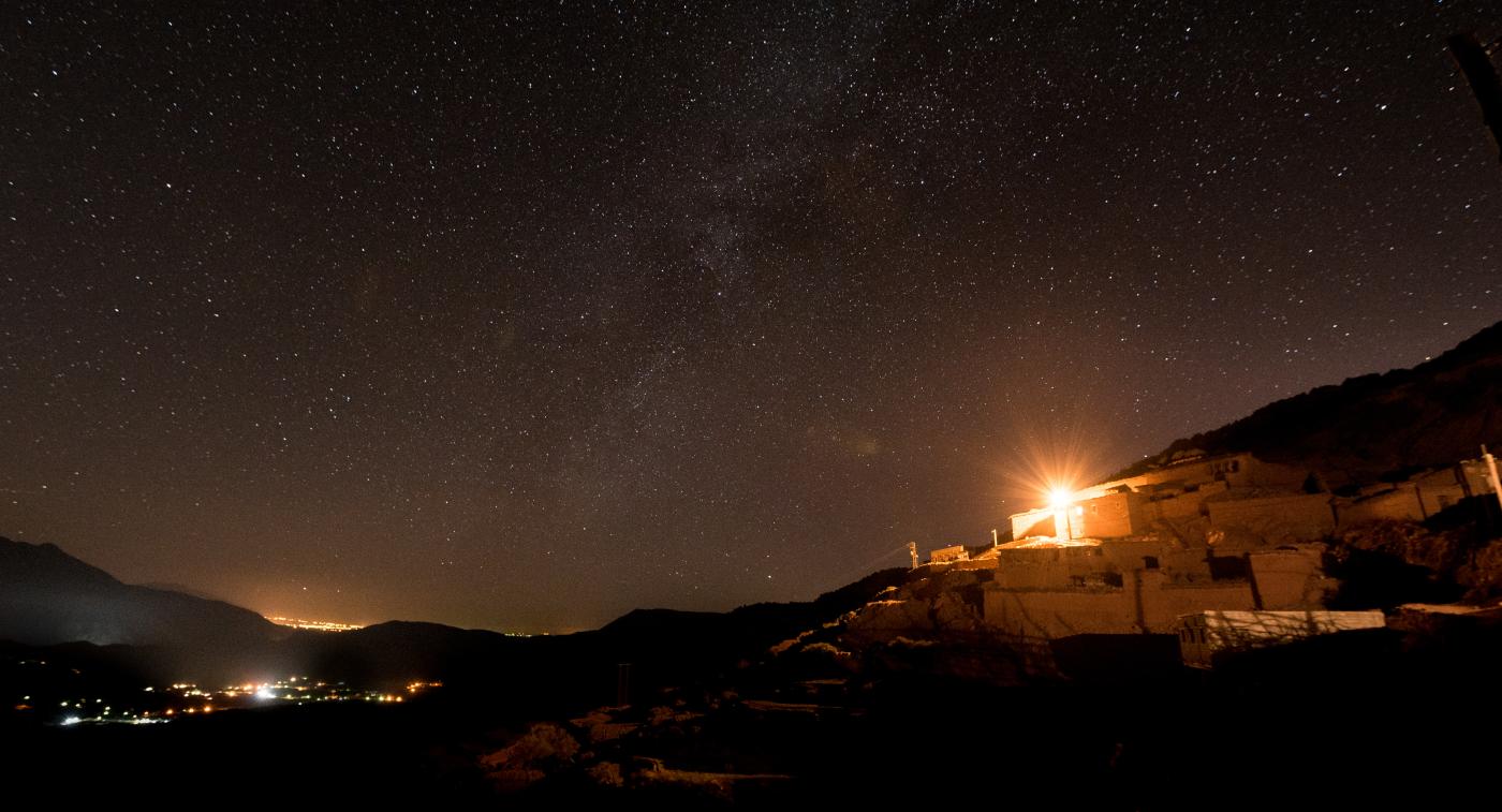 A Middle Eastern landscape under a night sky full of stars, with a bright lightshining above a small house on the horizon.