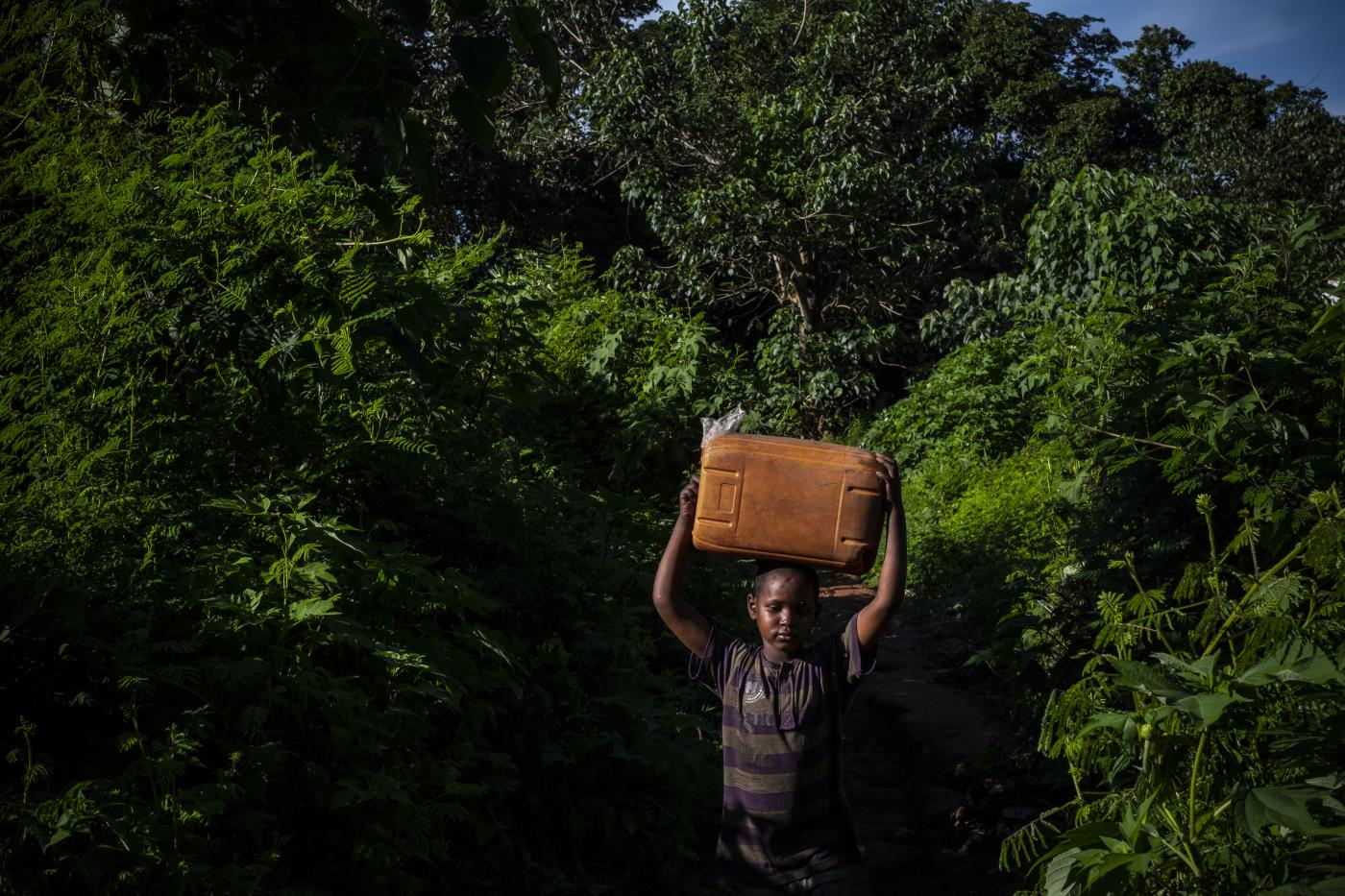 A child carrying a big plastic canister stepping out from a lush forest.