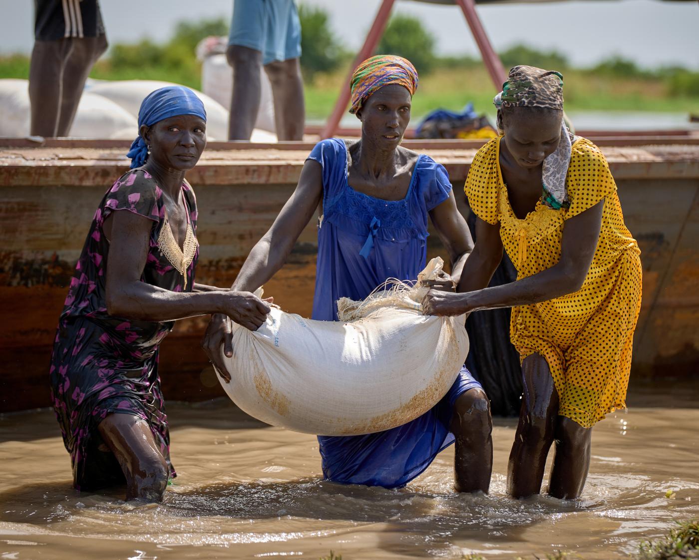 African women walk in water carrying a white sack containing food. 