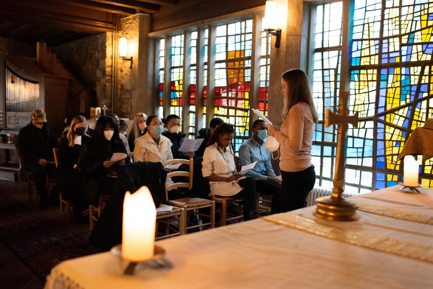 People gathered in a stained glass chapel room, most of them seated, one young woman reading from a paper.