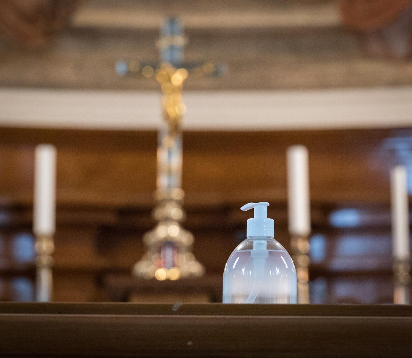 Bottle of hand sanitizer sits on wooden table in the foreground, in front of a blurred crucifix in the background.