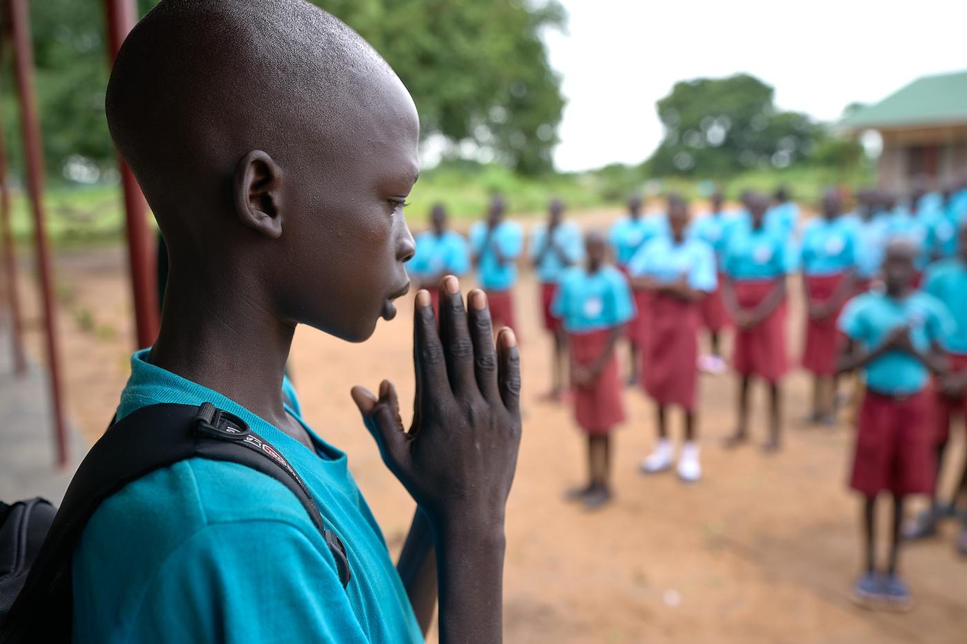 A young boy holds the palms of his hands together by his face in prayer. Other school children look on. 