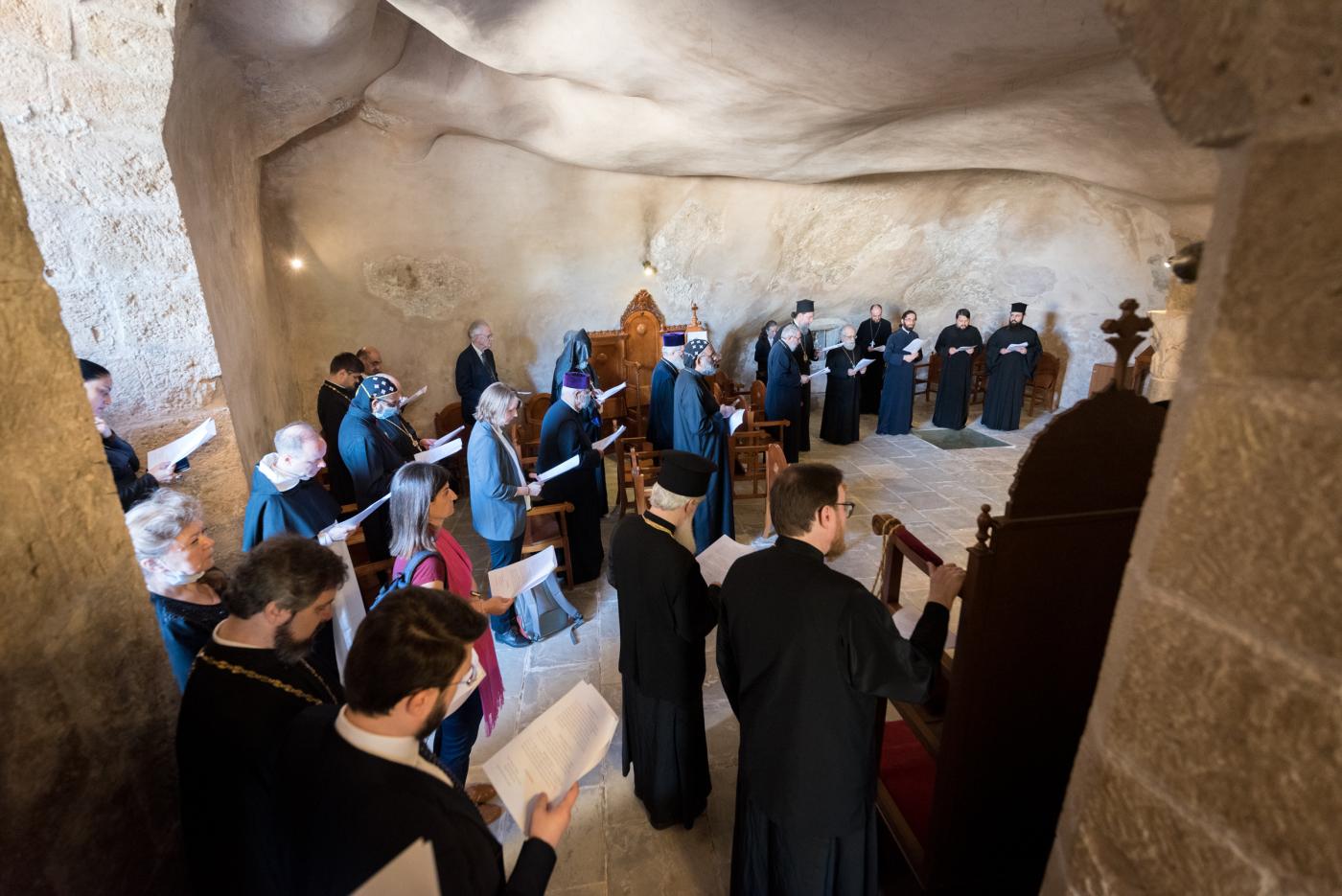 Group of people of different ages, many of them clergy wearing black religious garb, gather inside a stone wall chapel to pray. 