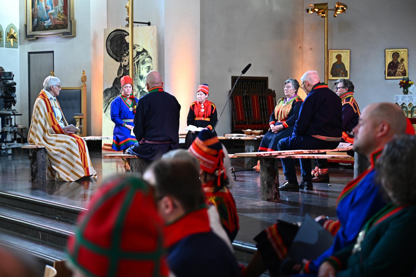 Group of people, some dressed in religious garb and some in traditional Sami indigenous clothing, gathered in a circle in a church building.