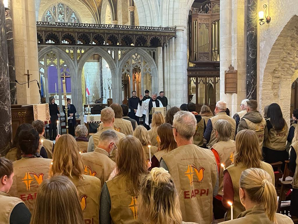 People wearing yellow vests reading 'EAPPI' gathered in a church where a pastor is giving them a blessing. 