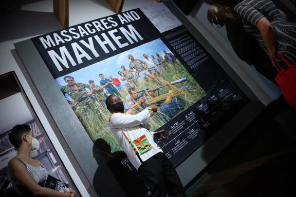 African man wearing a black medical face mask stands in front of a poster at a museum. The poster reads 'Massacres and mayhem', shoing images of past war times in South Africa. 
