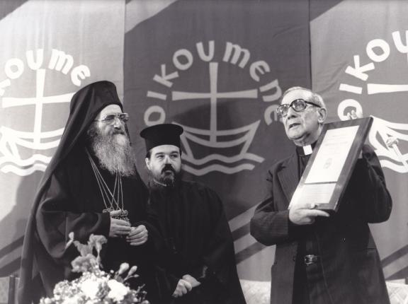 Man holding diploma with 'oikoumene' symbols in the background, and two other men looking on. 