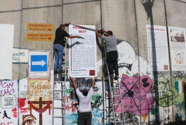 Two people stand on ladders in order to glue a poster onto a tall concrete wall, supported by a third person standing on the ground. 