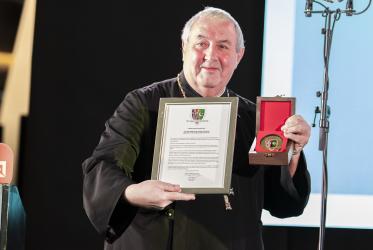 A man smiles while posing for a photo holding a framed diploma and a badge given to him as an award.