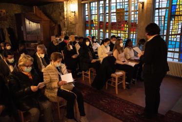 People sitting on chairs praying in a chapel with stained glass in the background. 