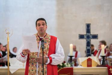 Man dressed in religious garb pictured in a church standing at a microphone speaking. 